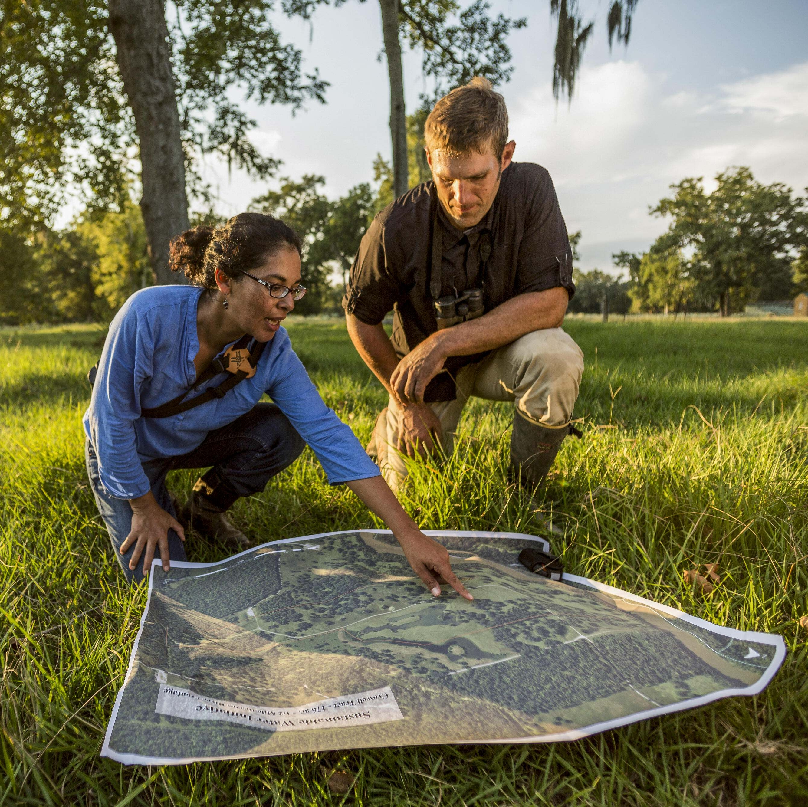 A woman and man crouch in green grass reviewing a map.